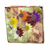 A set of four square coasters with a colorful floral pattern, featuring FleuroSun sunflowers, placed in a brown box with a camouflage-patterned background.