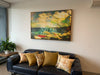 "Bathing in Nostalgia" archival canvas print by Durham J White, featuring Mother's Beach, Mornington, with beach huts, swimmers, shifting turquoise blues, light on the water, and a vibrant splash of red, shown box framed in a modern apartment styled with Durham J White Belgian linen cushions in sand and mustard tones.