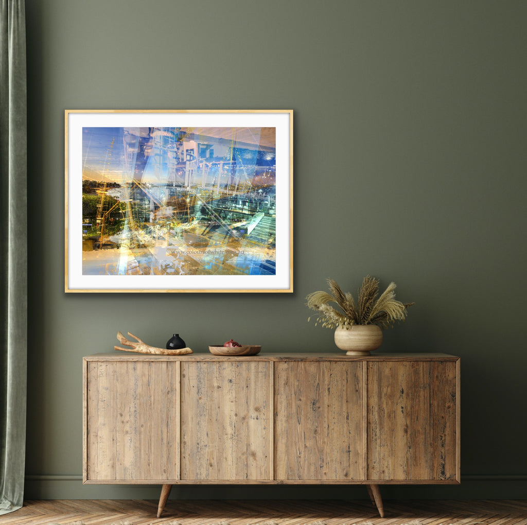 “Keeping Up a Pier Glance 2” glass-framed fine art print by Durham J White, featuring a layered photographic artwork of the Mornington waterfront in luminous pastel blues, subtle lemon highlights, and warm orange accents. Featured in a glass frame in a sage toned living or bedroom space.