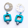 A pair of Elfy earrings featuring a silver-colored circular design with aqua squares, with visible earring hooks.