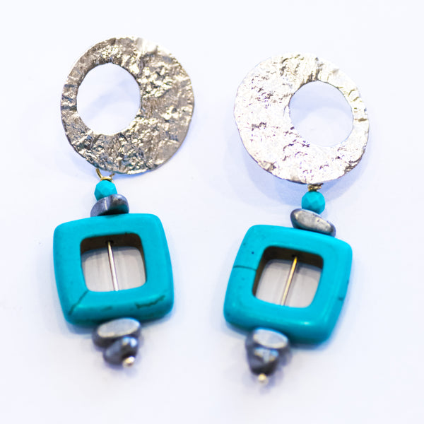 A pair of Elfy earrings featuring a silver-colored circular design with aqua squares, with visible earring hooks.