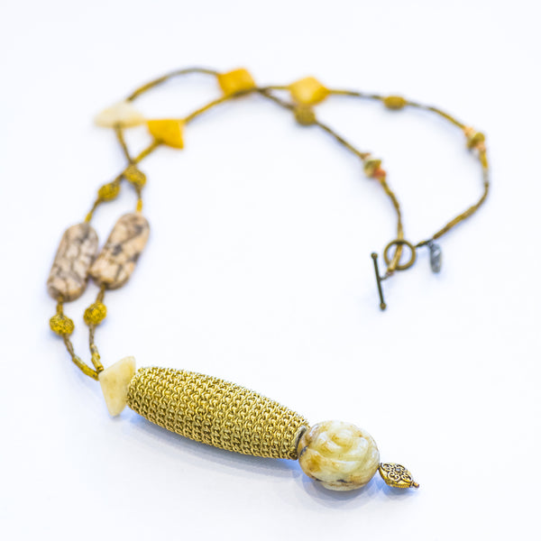 A long, golden necklace with a cylindrical feature and flower carving, displayed against a white background.