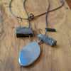 A long thin hematite strand necklace with a titanium plated feature pendant and two smaller raw cut gems, displayed on a wooden surface.